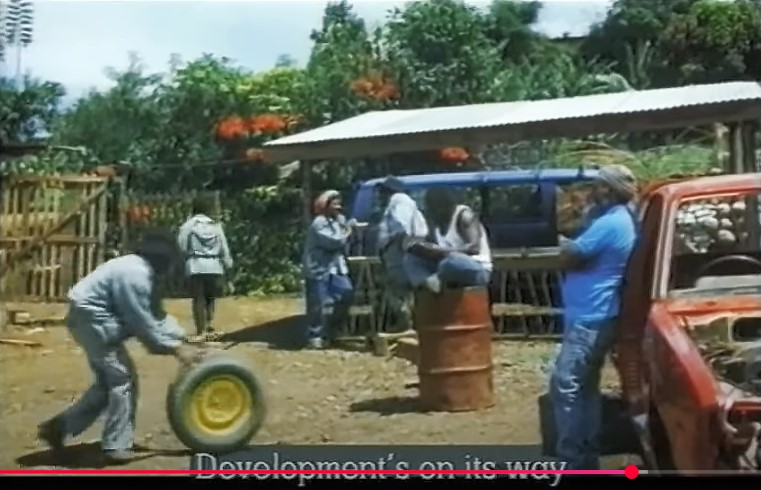 Un homme pousse devant lui une roue de secours alors que Papa (Leo Konga) attend devant son taxi rouge endommagé.