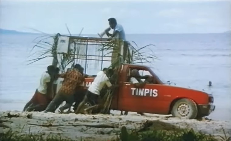 Peter Subek (Gerard Gabud) encourage ses hommes avec un haut-parleur pour qu'ils dépannent le taxi rouge Tinpis conduit par Papa (Leo Konga).