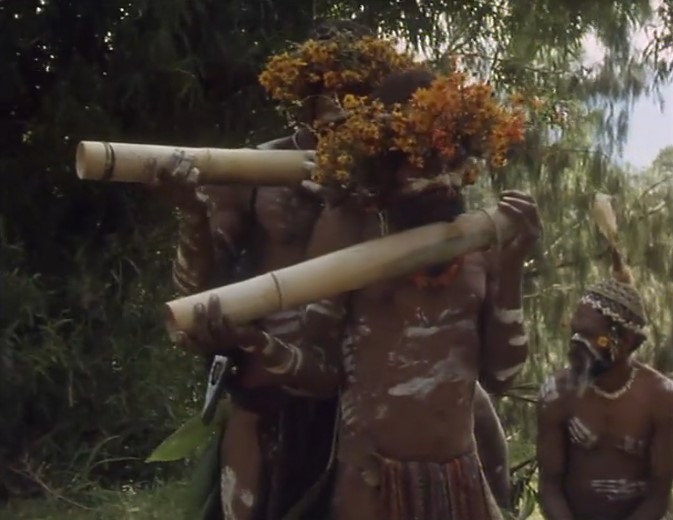 Deux Papous de Chimbu couronnés de fleurs jouent d'énormes flûtes tandis qu'un 3e les écoute.