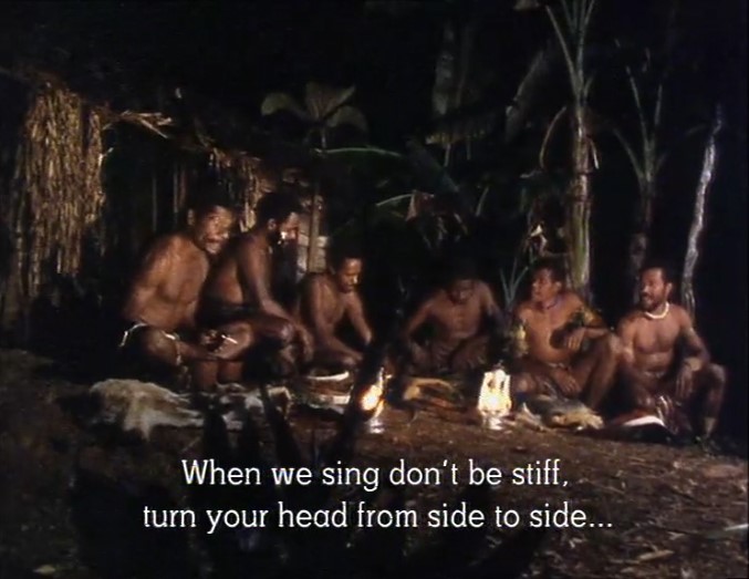 Un groupe de 6 jeunes Papous veillent la nuit en plein air autour d'une lampe pour préparer leurs chants. L'un d'eux les exhorte: "ne soyez pas raides en chantant, tournez vos têtes sur les côtés".