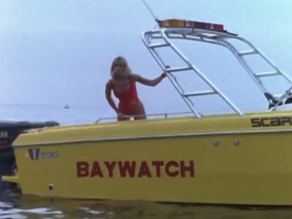 Julie (Tai Collins) en maillot de bain rouge se penche du bateau de secours Baywatch pour secourir une naufragée.