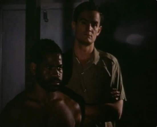 Le Papou T'Laupo (Justin Alu) comparaît au tribunal, gardé par un officier (Ric Chappel).