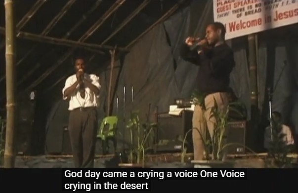 Deux prédicateurs papous exhortent la foule sur une estrade, devant une affiche proclamant "Bienvenue en Jésus". La légende dit en anglais: "le jour de Dieu venait, une voix criant dans le désert".