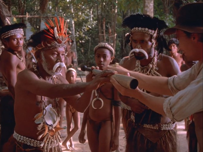 Le jeune Indiana Jones (Sean Patrick Flanery) donne une longue vue au chef (Tau Retu) d'une île de l'archipel Massim de Papouasie-Nouvelle-Guinée; le chaman (Sam Kaniyoisi) et le jeune Biok (Siloe Marum) les observent.