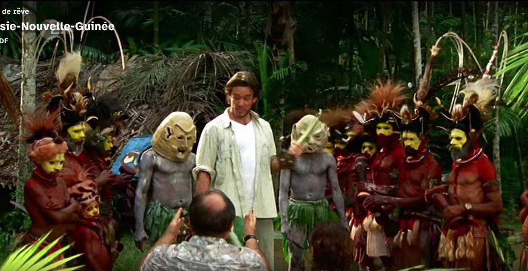 Le guide Joe (Jan Hartmann), entouré de deux guerriers masqués Asaros et de danseurs Hulis, s'approche de Dirk (Markus Majowski) et d'Erika (Manon Straché) assis au premier plan.