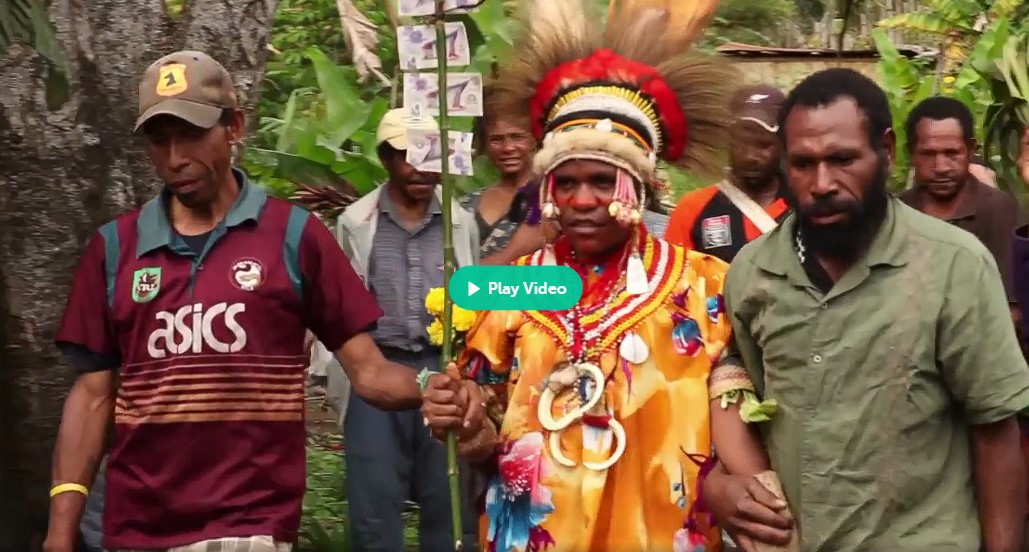 Aliko (Moslyn Moses), entourée de deux hommes de la tribu, est parée de coquillages et coiffée de plumes pour son mariage arrangé avec le vieux Kole. Elle tient à la main une perche avec des billets de banque reçus pour le paiement de son prix de fiancée.
